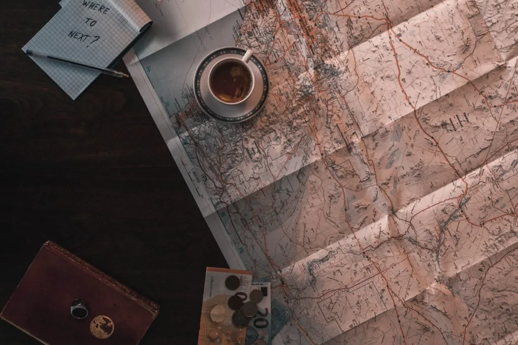 A map, coffee mug, notebook with "WHERE TO NEXT?" written, coins, and a camera on a wooden surface