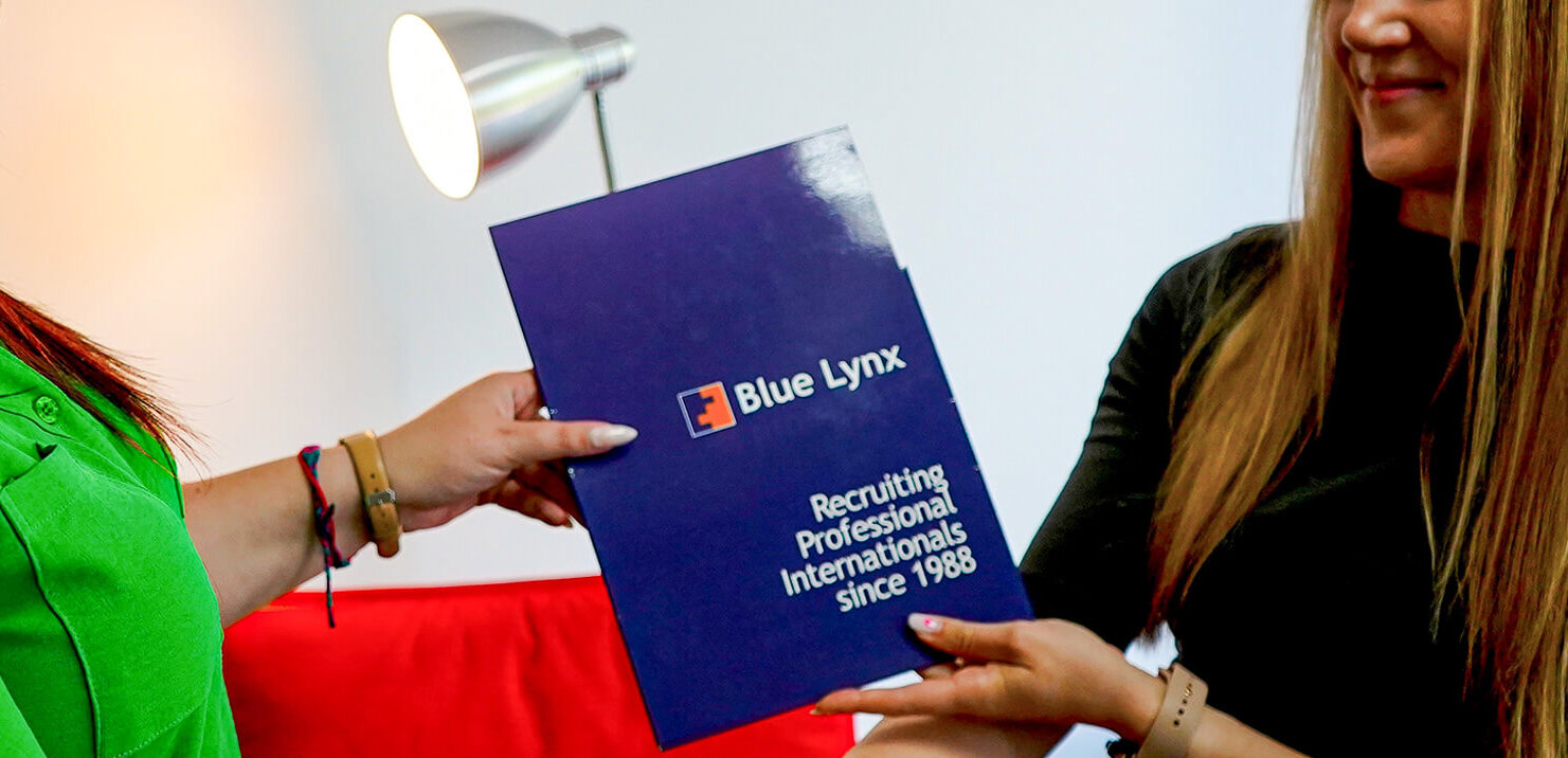 Two women face each other, each holding a blue book, engaged in a discussion or sharing information about recruitment services.